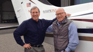 Two men smiling in front of a small airplane.