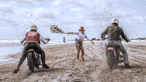 A woman waves a checkered flag at a muddy motorcycle race on a beach.