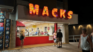 Neon sign reading 'MACKS' above a food stand with customers.