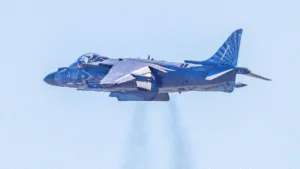 A blue military jet flying with exhaust smoke visible.